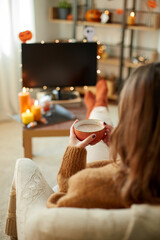 Poster - halloween, holidays and leisure concept - young woman watching tv and drinking hot chocolate with her feet on table at cozy home