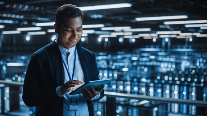 Wall Mural - Handsome Smiling IT Specialist Using Tablet Computer in Data Center. Succesful Businessman and e-Business Entrepreneur Overlooking Server Farm Cloud Computing Facility.