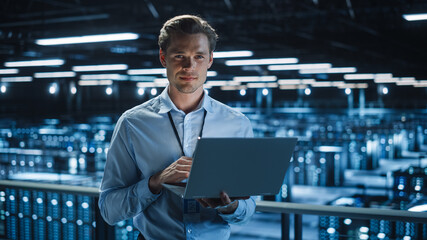 Wall Mural - Handsome Smiling on Camera IT Specialist Using Laptop Computer in Data Center. Succesful Businessman and e-Business Entrepreneur Overlooking Server Farm Cloud Computing Facility.