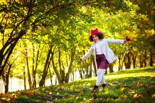 A Cute Little Mixed-race Girl In A Gray Coat, Skirt And Cap With A Bouquet Of Leaves In Her Hands Runs Through A Sunny Autumn Park. Copy Space.