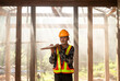 © somchai20162516 - Asian young professional carpenter with wooden planks and tools stand in his wooden workshop as workers work in the background of an old wooden house
