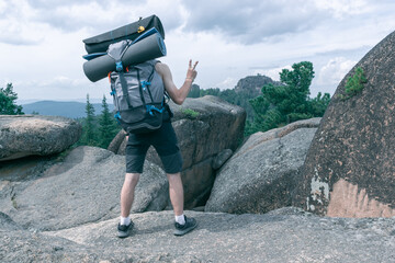 Back view of a guy with a backpack and camping equipment with an outstretched hand with the emotion of peace on the background of a beautiful landscape with rocks