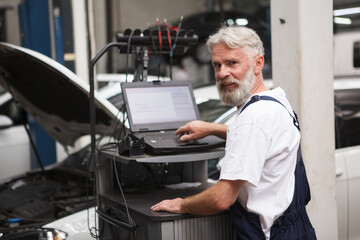 Wall Mural - Elderly car mechanic smiling to the camera while repairing an auto at the workshop, using computer