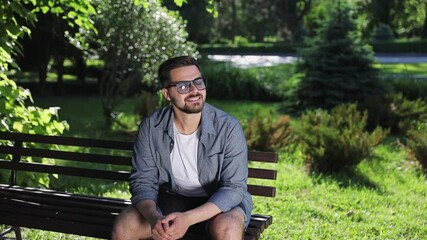 Wall Mural - Young smiley handsome stylish man 20s wears blue shirt glasses sit on wooden bench wait girlfriend rest relax in spring green city park sunshine day outdoors on nature. Urban lifestyle leisure concept