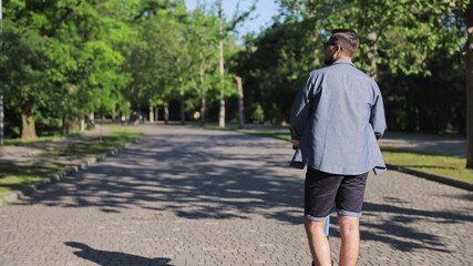 Wall Mural - Back rear view young man 20s wears blue shirt shorts glasses riding electric scooter go down alley rest relax in spring green city park sunshine lawn outside on nature. Urban leisure lifestyle concept