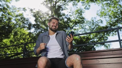 Wall Mural - Happy fun young man 20s wears blue shirt shorts earphones sit on bench use mobile cell phone enjoy listen to music rest relax in spring green city park outdoor nature Urban leisure lifestyle concept