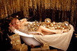 Christmas Woman portrait over gold xmas gifts and balls in bathtub. Beauty fashion model girl with make up, wavy hair and jewellery, posing in golden gems dress over bright bokeh background.