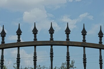 a row  iron sharp rods on a black  wall of a metal fence against a background of blue sky and white clouds