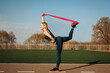 © Natalia - Sporty woman is stretching her back and leg using expander, resistance band outdoors at the stadium.