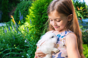 A cute girl is holding a white baby bunny on her chest. A dwarf decorative rabbit in the hands of a blonde girl. The concept of friendship between a child and a rodent pet.