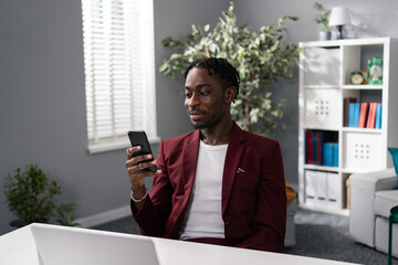 Wall Mural - Dark-skinned employee, boss manager, businessman dressed in maroon jacket American African man sits in chair at desk with phone in hand, smiles softly, reads, browses social media, break at work, rest