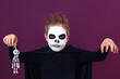 © Natali Illar - Little boy in halloween costume holds skeleton and scares the camera on purple background.