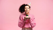 Young Woman In Party Cap Blowing Candle On Birthday Cupcake Isolated On Pink