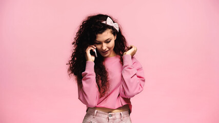 pretty and positive woman touching hair while talking on cellphone isolated on pink