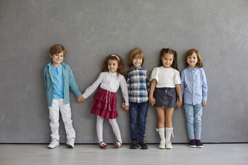 Portrait of cute and happy little boys and girls in casual clothes standing against the gray wall. Beautiful smiling and funny kids standing in a row holding hands. Childhood concept.