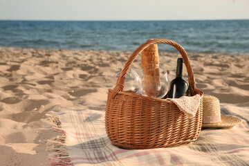 Poster - Blanket with bread, wine and glasses in basket for picnic near straw hat on sandy beach