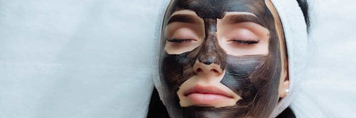 Wall Mural - banner . cosmetologist applying black mask on pretty woman face wearing black gloves, gorgeous woman in spa having facial procedures