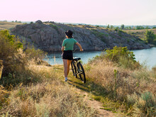 Girl In Green T-shirt With A Bike In The Field, Against The Backdrop Of Rocks, River, City