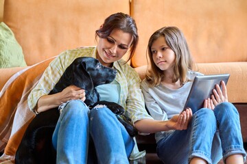 Mother daughter child and labrador puppy dog sitting together near sofa looking in digital tablet