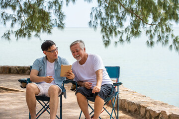 Happy Asian family handsome man teach senior father using digital tablet for video call or online shopping on the beach in summer day. Father and son enjoy and having fun together in holiday vacation