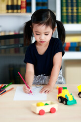 Little pretty pigtails hairstyle preschooler kindergarten girl sitting on chair drawing sketching painting cartoon with colored pencils on paper on table in living room at home in front bookshelf