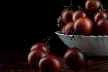 A lot of ripe juicy tomatoes in a white plate and next to it on a black background. Black tomatoes are cumato.