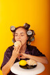 © Marina Chernivetskay - brunette girl greedily bites croissant on a yellow background with curlers on her head holding a croissant in her hands