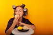 © Marina Chernivetskay - brunette girl greedily bites croissant on a yellow background with curlers on her head holding a croissant in her hands