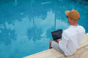 Young man wearing white linen shirt working on laptop by the pool, dipping his feet in a water. Male travel blogger typing on keyboard. Perks of being a freelancer. Close up, copy space, background.