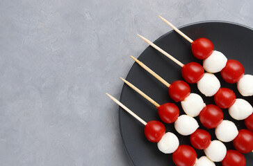 Wall Mural - Cold appetizer on wooden skewers: red cherry tomatoes and balls of mozzarella cheese on a black plate against a gray stone background. Copy space
