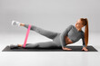Athletic girl doing leg raises exercise for glute with resistance band on gray background. Fitness woman working out