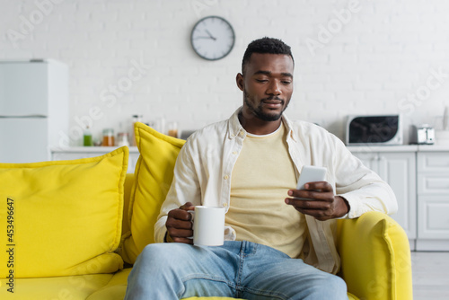 young african american man using smartphone while sitting on couch with cup