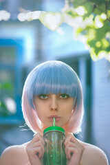 Wall Mural - Beautiful young woman with big eyes, makeup and colored pink and blue wig is drinking lemonade from the cactus shape mug in the summer evening backlight. Close up, selective focus