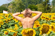 Shirtless woman with her back turned, emerging from a field of sunflowers with her arms and hands held high in the air.