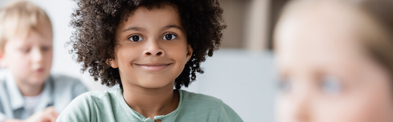 Wall Mural - happy african american boy smiling at camera near blurred classmates, banner