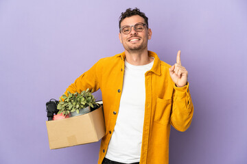 Young caucasian man making a move while picking up a box full of things isolated on purple background showing and lifting a finger in sign of the best