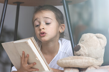 Poster - Little girl reads a book with a teddy bear.