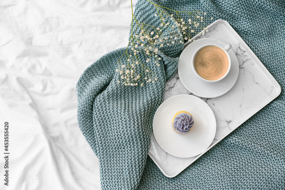 Tray with cup of coffee, cupcake and flowers on bed