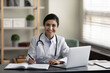© fizkes - Portrait of young smiling confident indian ethnicity female doctor general practitioner therapist in medical uniform working on computer, making records in modern clinic office room sitting at table.