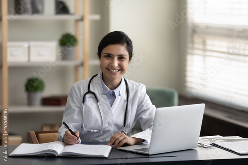 Portrait of young smiling confident indian ethnicity female doctor general practitioner therapist in medical uniform working on computer, making records in modern clinic office room sitting at table.