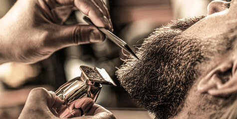 Wall Mural - Barber works with a beard clipper. Hipster client getting haircut. Hands of a hairdresser with a beard clipper, closeup. Bearded man in barbershop. Man visiting hairstylist in barbershop