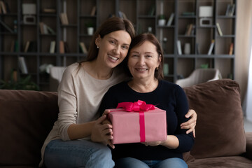 Canvas Print - Portrait of smiling affectionate young woman cuddling sincere joyful elder mature retired mother holding wrapped box with present gift in hands, happy birthday or special event celebration concept.