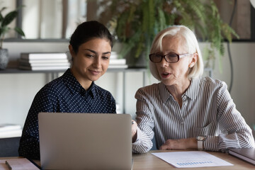 Serious mature businesswoman mentor coach in glasses teaching Indian woman intern student, using laptop, pointing at screen, two diverse colleagues working on online project together, discussing