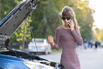 Wall Mural - Angry female driver talking on cell phone with assistance service worker standing near a broken car with popped up hood on road side having trouble with her vehicle.