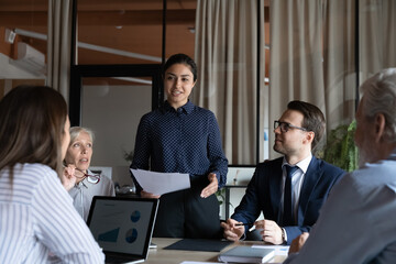 Wall Mural - Smiling young Indian businesswoman mentor executive speaking at corporate meeting, explaining project strategy, presenting statistics, diverse colleagues coworkers employees involved in briefing