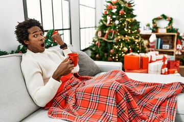 Sticker - Young african american man sitting on the sofa drinking coffee by christmas tree surprised pointing with hand finger to the side, open mouth amazed expression.