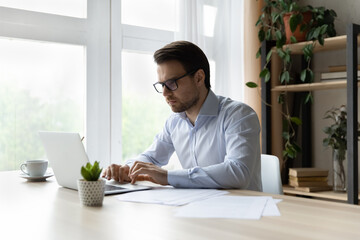 Canvas Print - Serious millennial businessman working at laptop in office. Focused young man in glasses, employee, lawyer using computer at workplace, typing, chatting online, watching virtual presentation