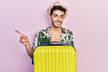 Wall Mural - Young hispanic man wearing summer style and holding cabin bag smiling and looking at the camera pointing with two hands and fingers to the side.