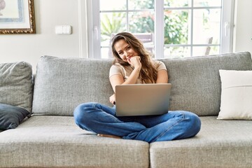 Beautiful young brunette woman sitting on the sofa using computer laptop at home looking confident at the camera smiling with crossed arms and hand raised on chin. thinking positive.