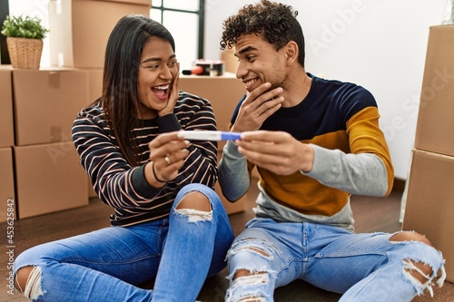 Young latin couple surprised holding pregnancy test sitting on the floor at new home.